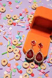 Red teardrop earrings in an orange jewelry box with colorful sprinkles and cereal on a purple background