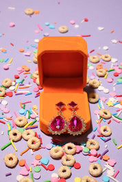 Earrings with pink gemstones in an open orange jewelry box surrounded by colorful sprinkles and donut-shaped cookies on a purple background.