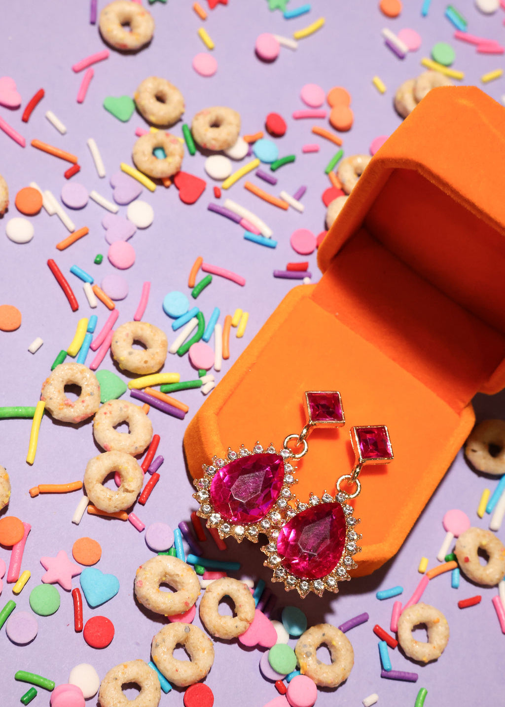 Pink gemstone earrings in an orange jewelry box with colorful sprinkles and cookies on a purple background