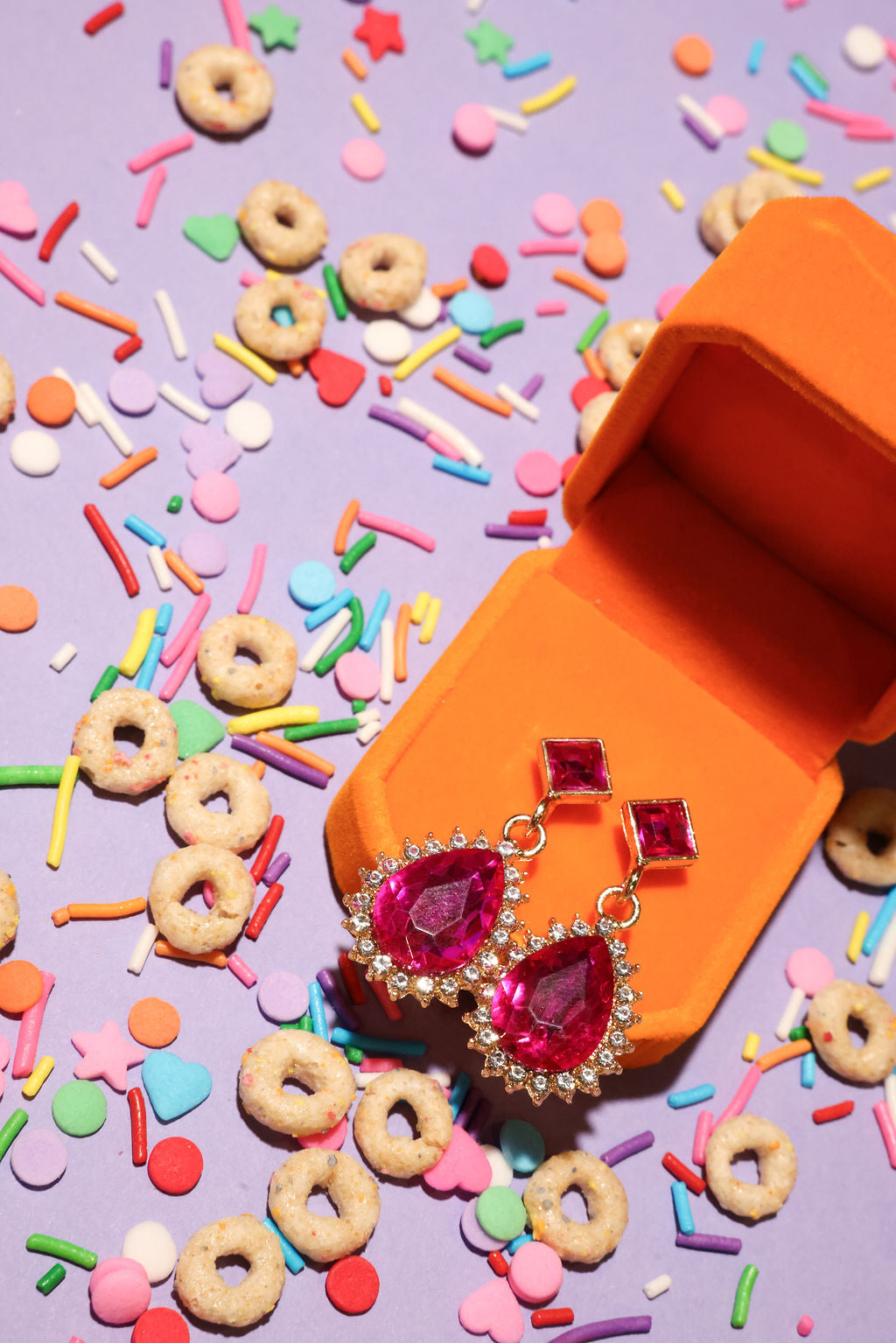 Pink gemstone earrings in an orange jewelry box with colorful sprinkles and cookies on a purple background