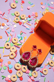 Pink gemstone earrings in an orange jewelry box with colorful sprinkles and cookies on a purple background