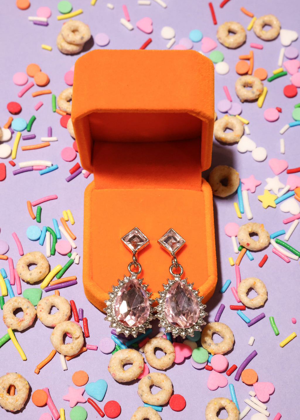Open orange jewelry box with sparkling earrings on a colorful cereal and sprinkles background