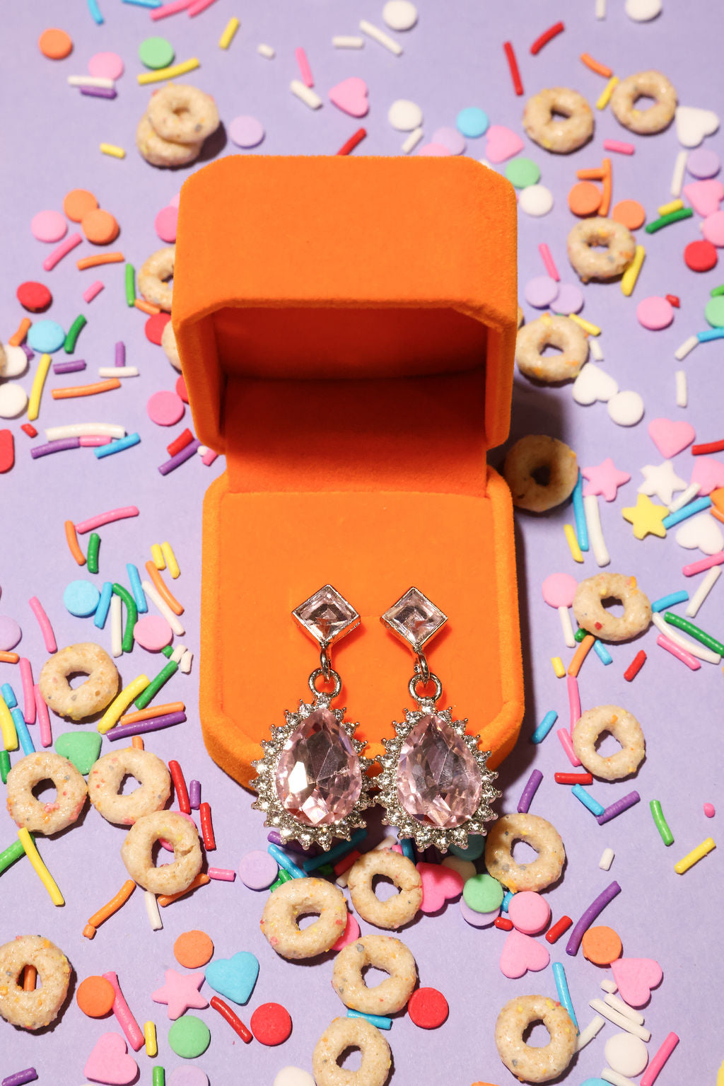 Open orange jewelry box with sparkling earrings on a colorful cereal and sprinkles background