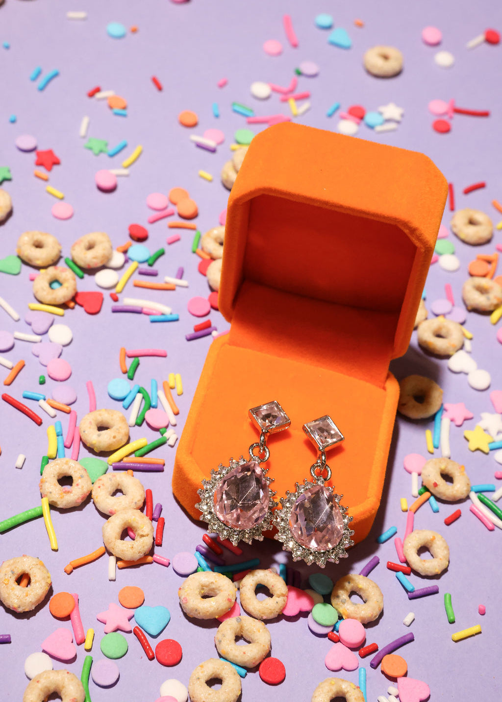 Silver earrings in an orange jewelry box surrounded by colorful sprinkles and donut-shaped candies on a purple background.