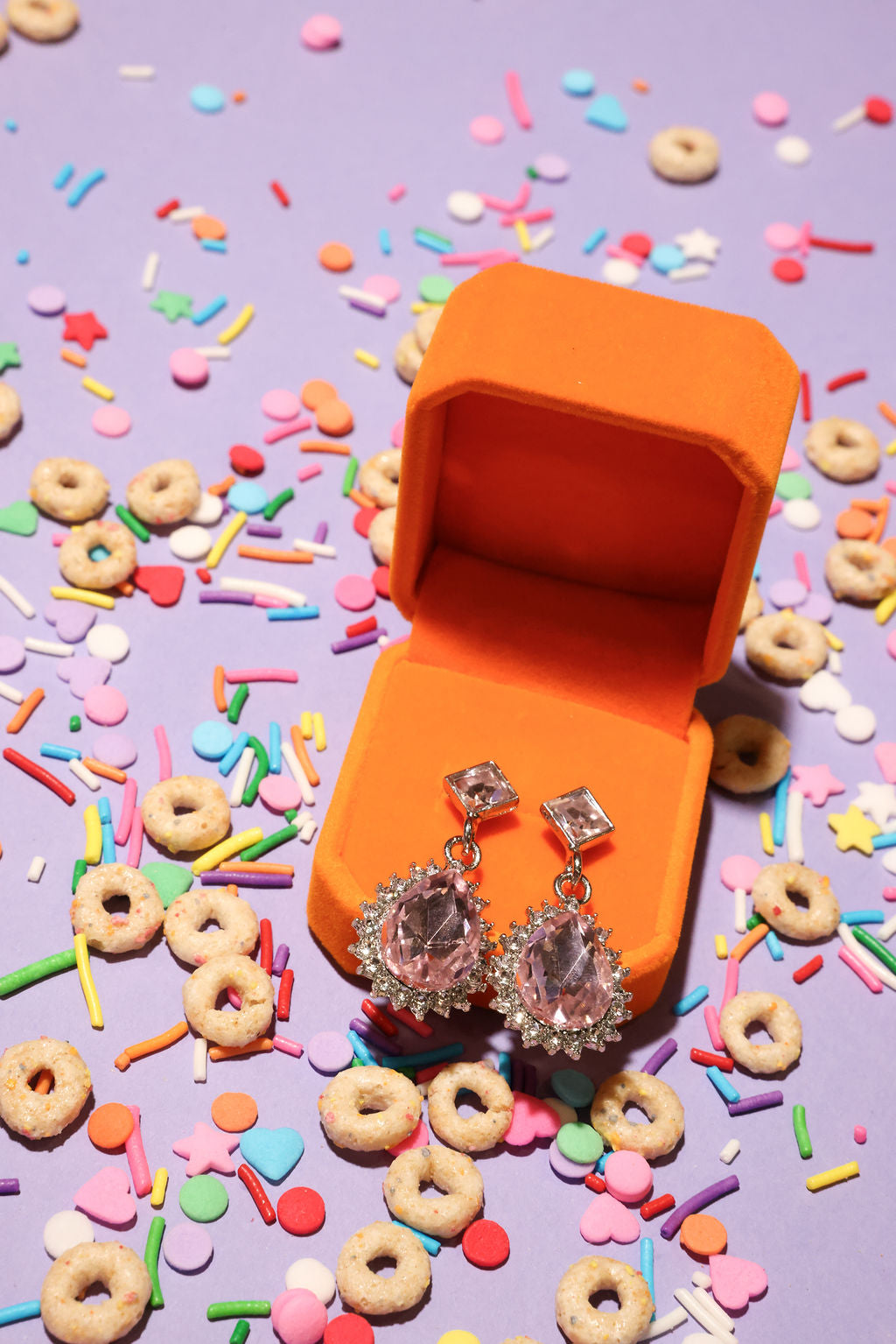 Silver earrings in an orange jewelry box surrounded by colorful sprinkles and donut-shaped candies on a purple background.