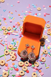 Silver earrings in an orange jewelry box surrounded by colorful sprinkles and donut-shaped candies on a purple background.
