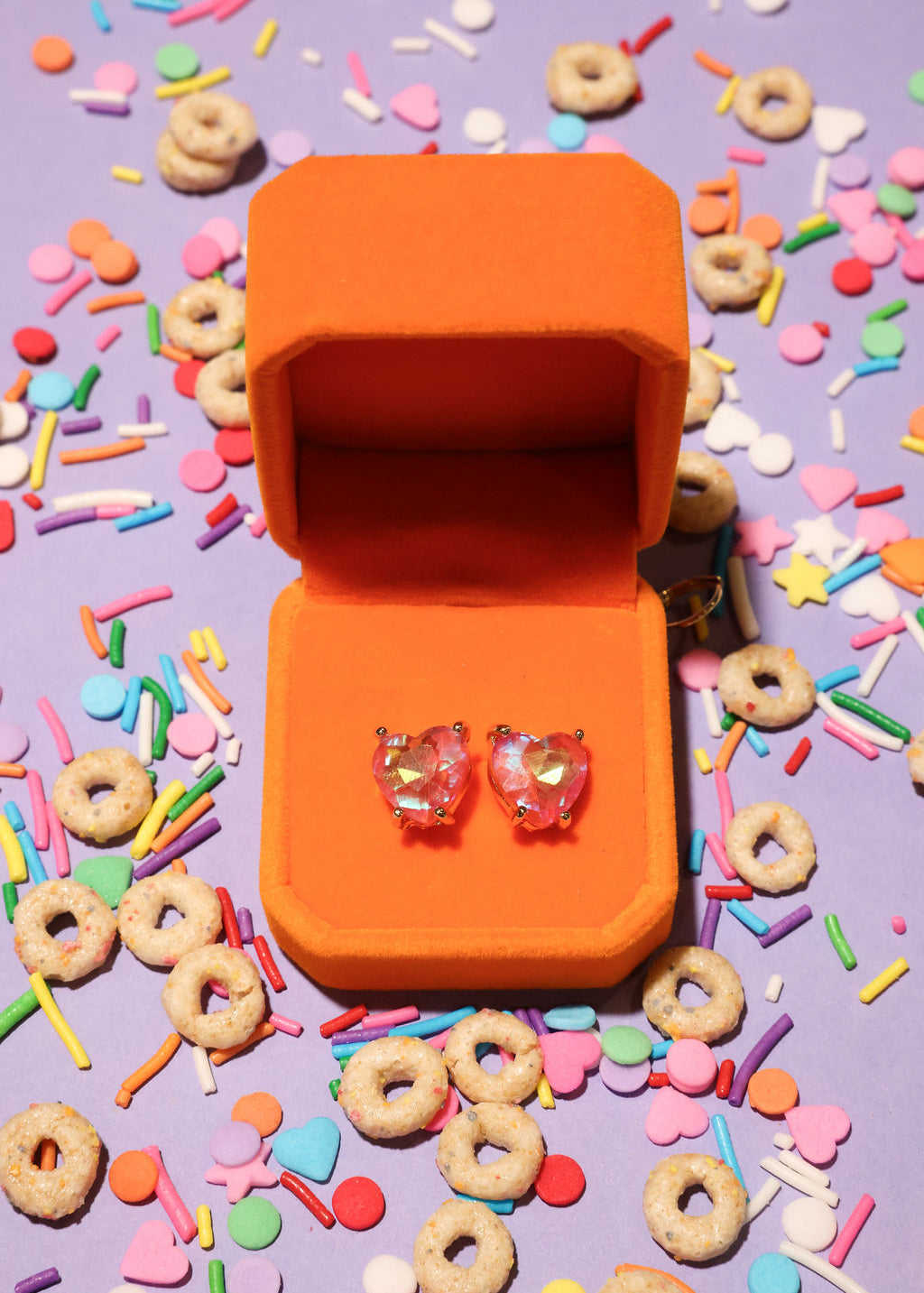 Heart-shaped earrings in an orange box surrounded by colorful sprinkles and donuts on a purple background