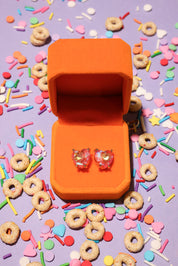Heart-shaped earrings in an orange box surrounded by colorful sprinkles and donuts on a purple background