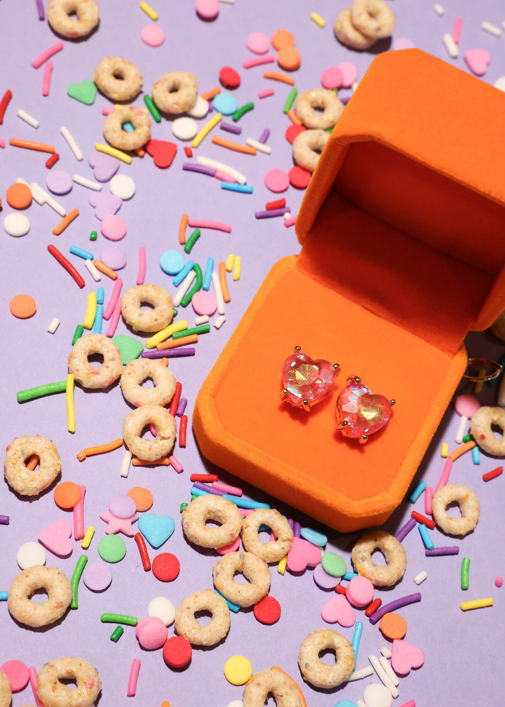 Orange jewelry box with heart-shaped earrings on a colorful sprinkles and cereal background