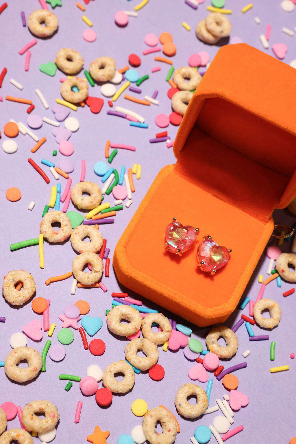 Orange jewelry box with heart-shaped earrings on a colorful sprinkles and cereal background