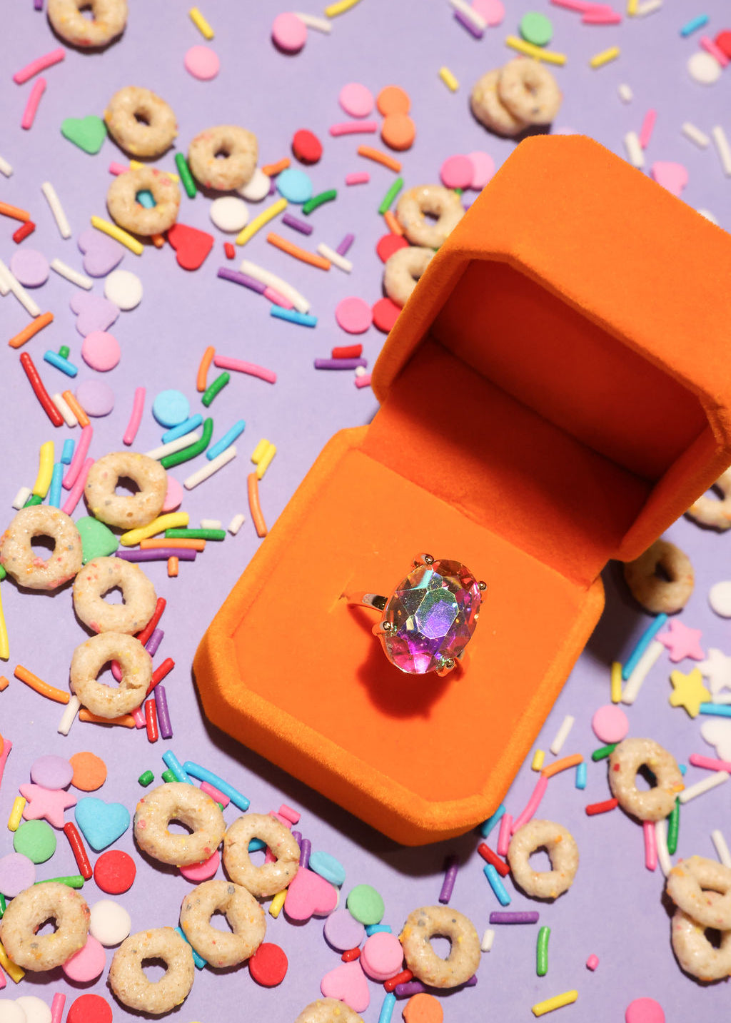 Orange jewelry box with a ring on a purple background with colorful sprinkles and donut-shaped cereal