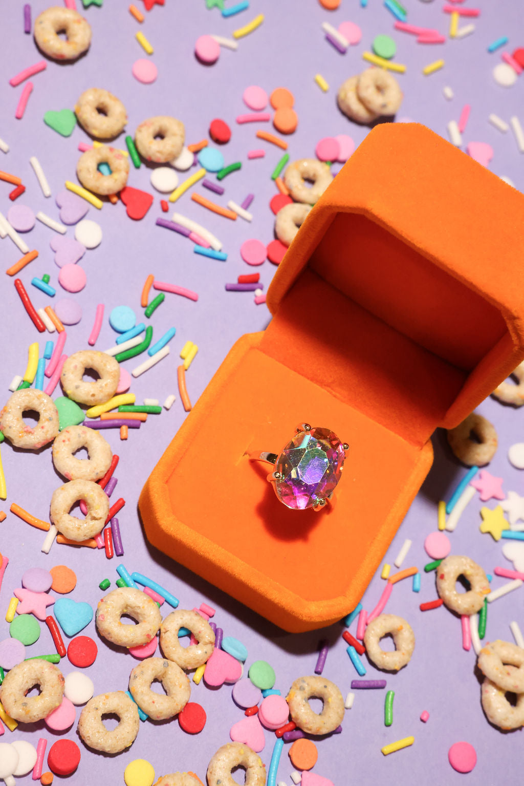 Orange jewelry box with a ring on a purple background with colorful sprinkles and donut-shaped cereal
