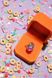 Orange jewelry box with a ring on a purple background with colorful sprinkles and donut-shaped cereal