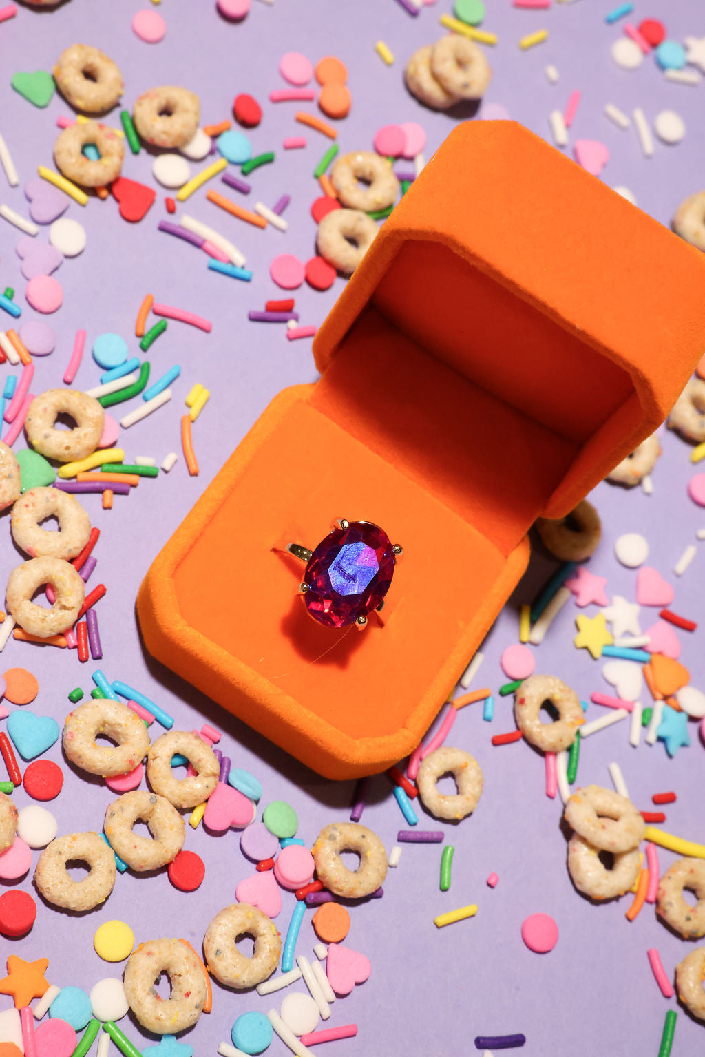 Orange jewelry box with a ring on a colorful background of sprinkles and donut-shaped cereal