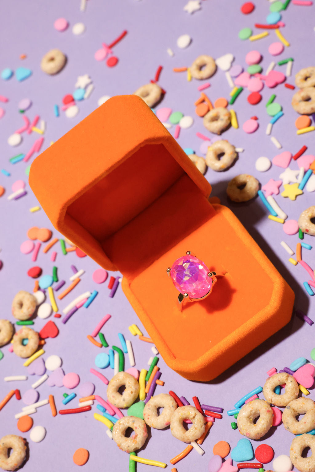 Orange jewelry box with a pink ring on a colorful background with sprinkles and cereal