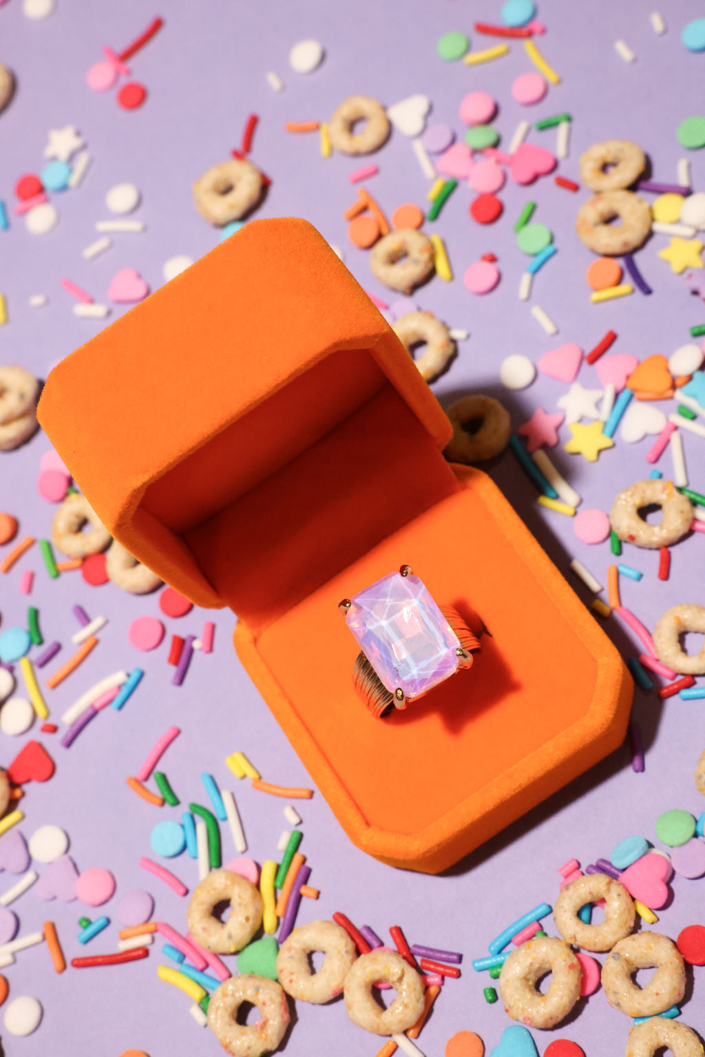 Orange jewelry box with a ring on a purple background with colorful cereal pieces and sprinkles
