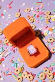 Orange jewelry box with a ring on a purple background with colorful cereal pieces and sprinkles