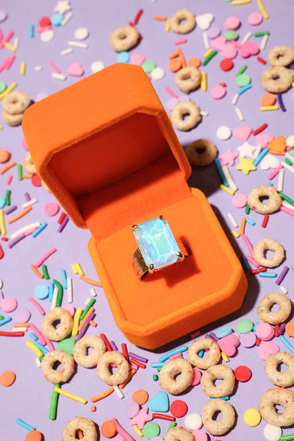 Orange ring box with a ring featuring a blue gemstone on a colorful sprinkles and cereal background