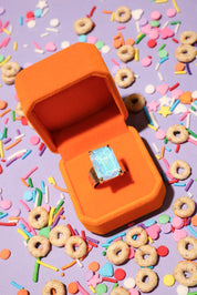 Orange ring box with a ring featuring a blue gemstone on a colorful sprinkles and cereal background