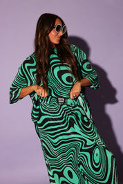 Person wearing a green and black patterned outfit with sunglasses on a purple background, frontal view