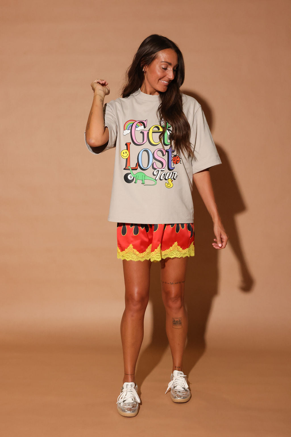 Woman wearing a 'Get Lost' t-shirt and colorful shorts on a beige background, frontal view