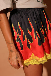 Model wearing a black, red, and yellow flame-patterned shorties with lace trim on a brown background, close up side view