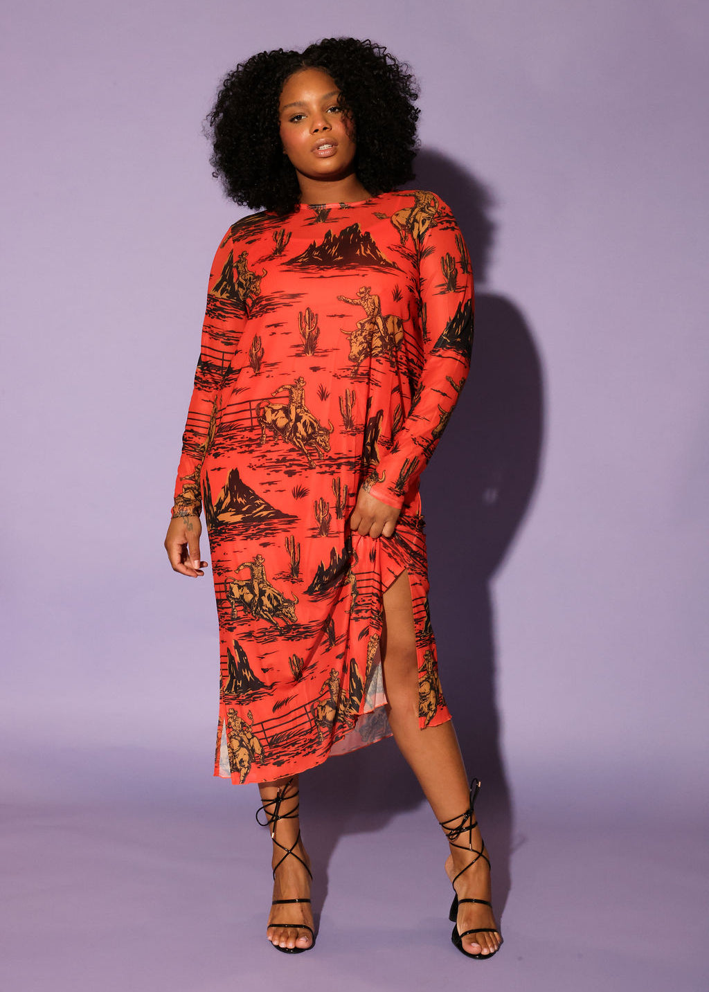 A model with curly black hair in a studio setting against a solid lavender background, wearing a long-sleeve dress in a vibrant orange-red color with a black graphic print, frontal view