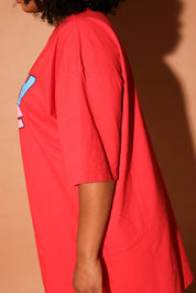 Person wearing a red oversized shirt with colorful design on a brown background, side view