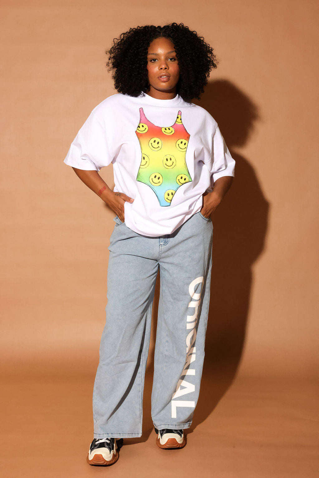 Model wearing a white t-shirt with a colorful swimsuit graphic and light blue jeans on a beige background, frontal view