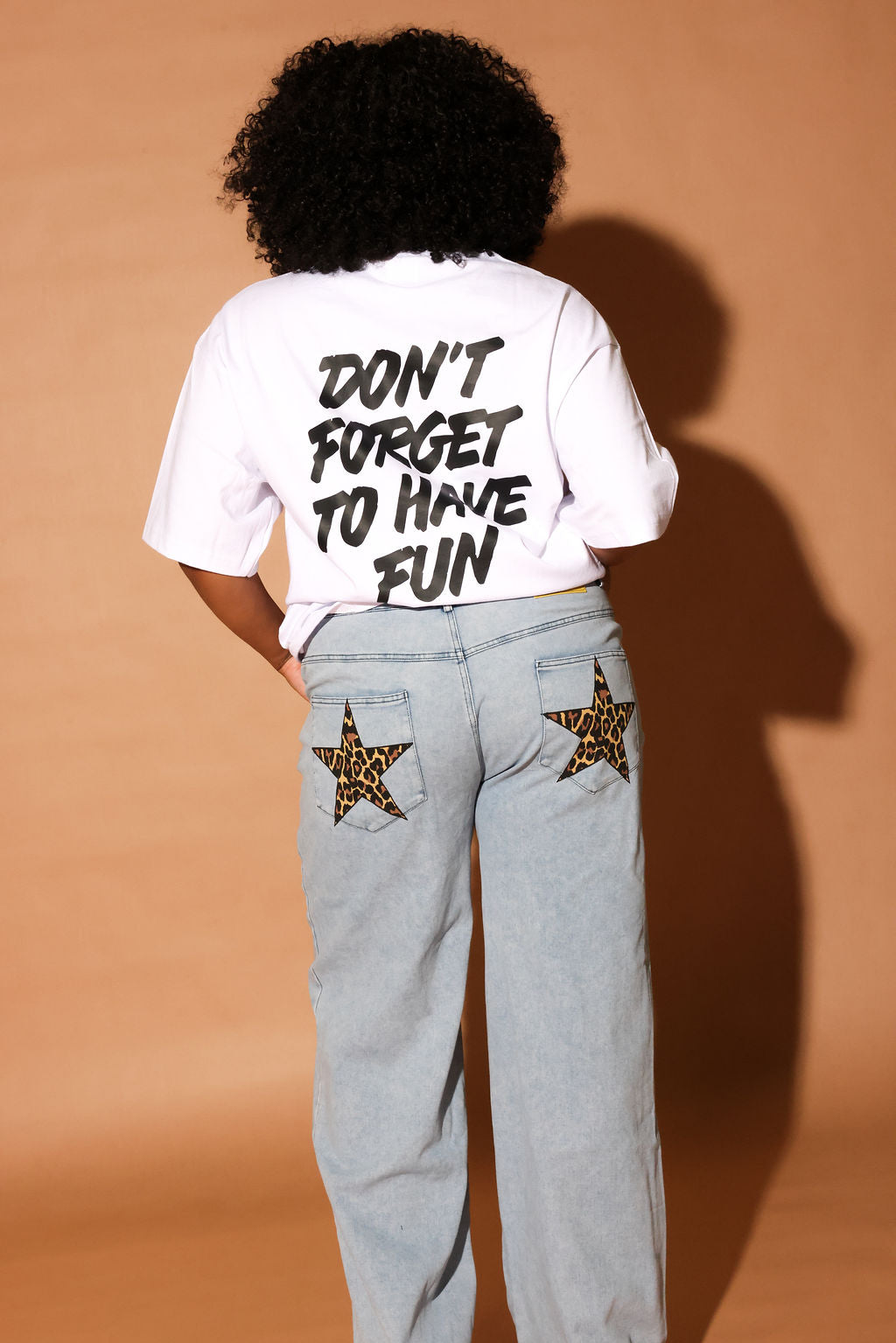 Model wearing a white t-shirt with text and light blue jeans with star designs on a brown background, back view