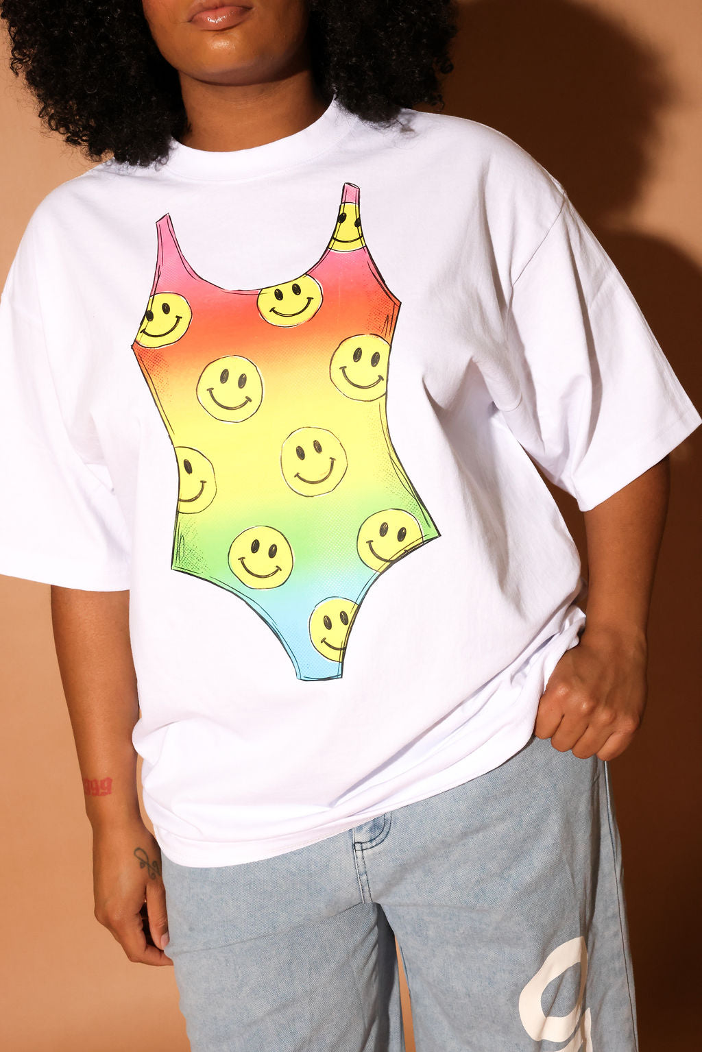 Model wearing a white t-shirt with a colorful swimsuit graphic and light blue jeans on a beige background, close up frontal view