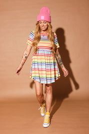 Woman wearing a colorful striped short dress and pink beanie against a brown background, frontal view