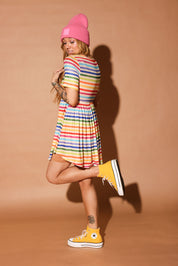Woman wearing a colorful striped short dress and pink beanie against a brown background, side view