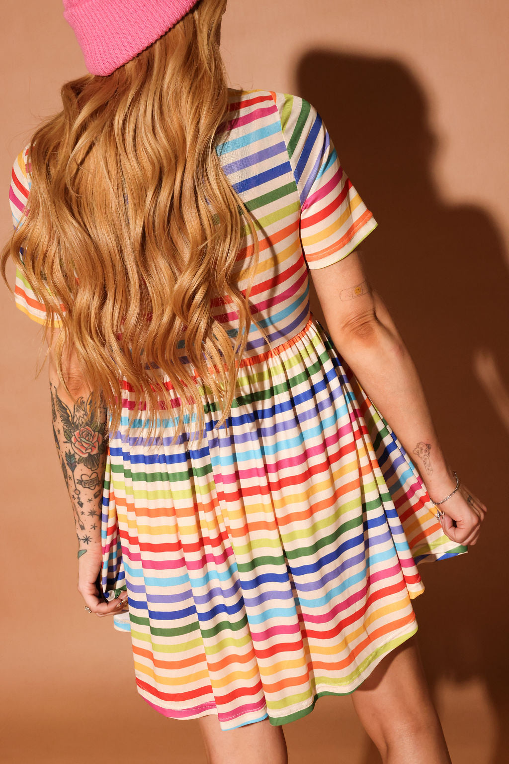 Woman wearing a colorful striped short dress and pink beanie against a brown background, back view