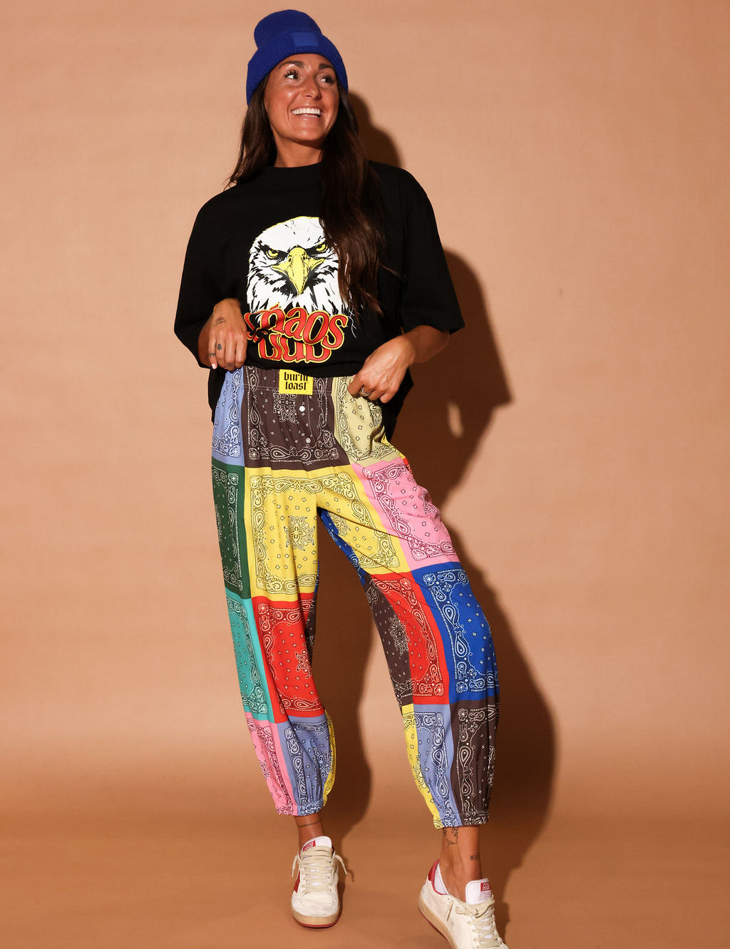 Person wearing a black t-shirt with an eagle graphic and colorful bandana pants on a beige background, frontal view
