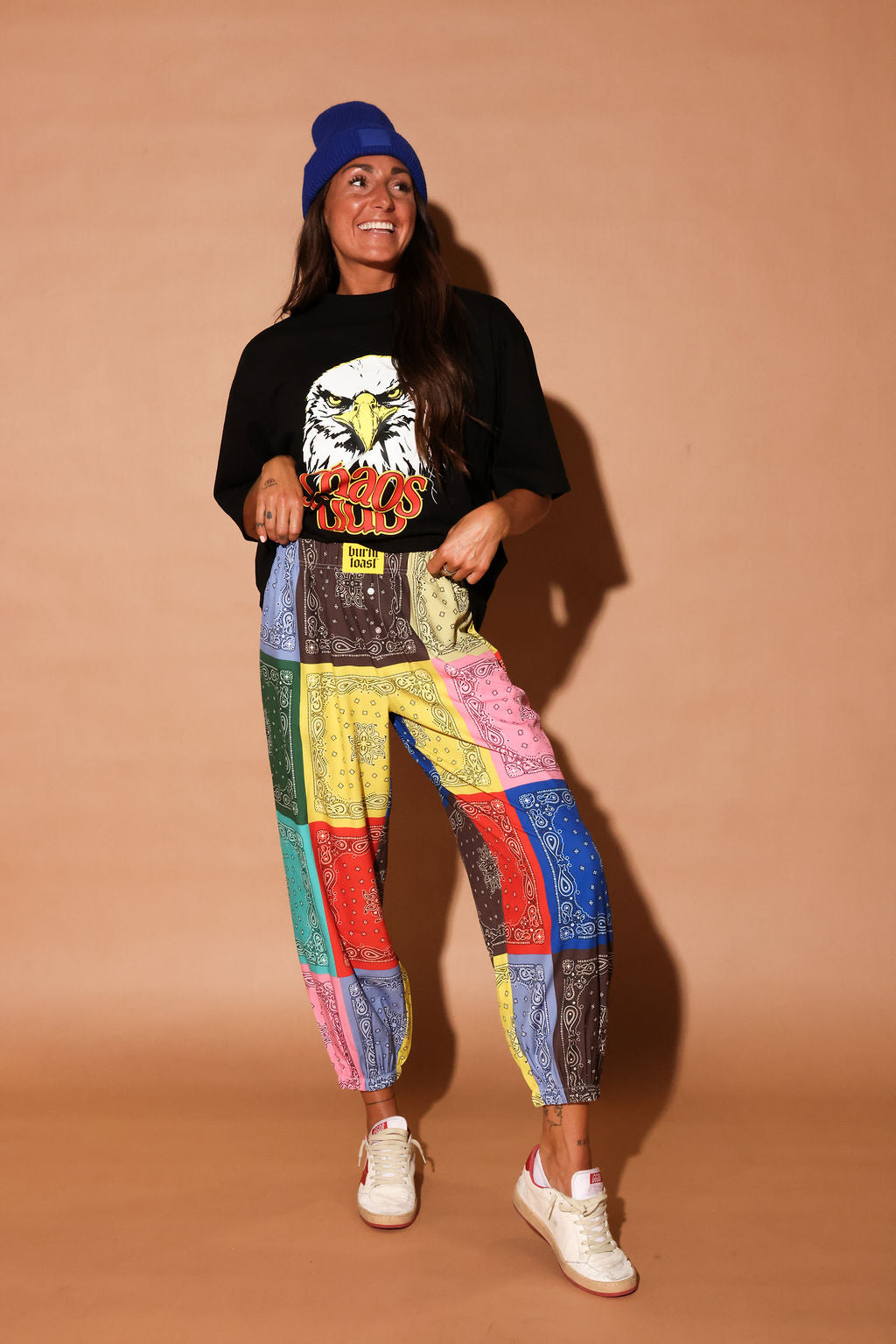 Person wearing a black t-shirt with an eagle graphic and colorful bandana pants on a beige background, frontal view