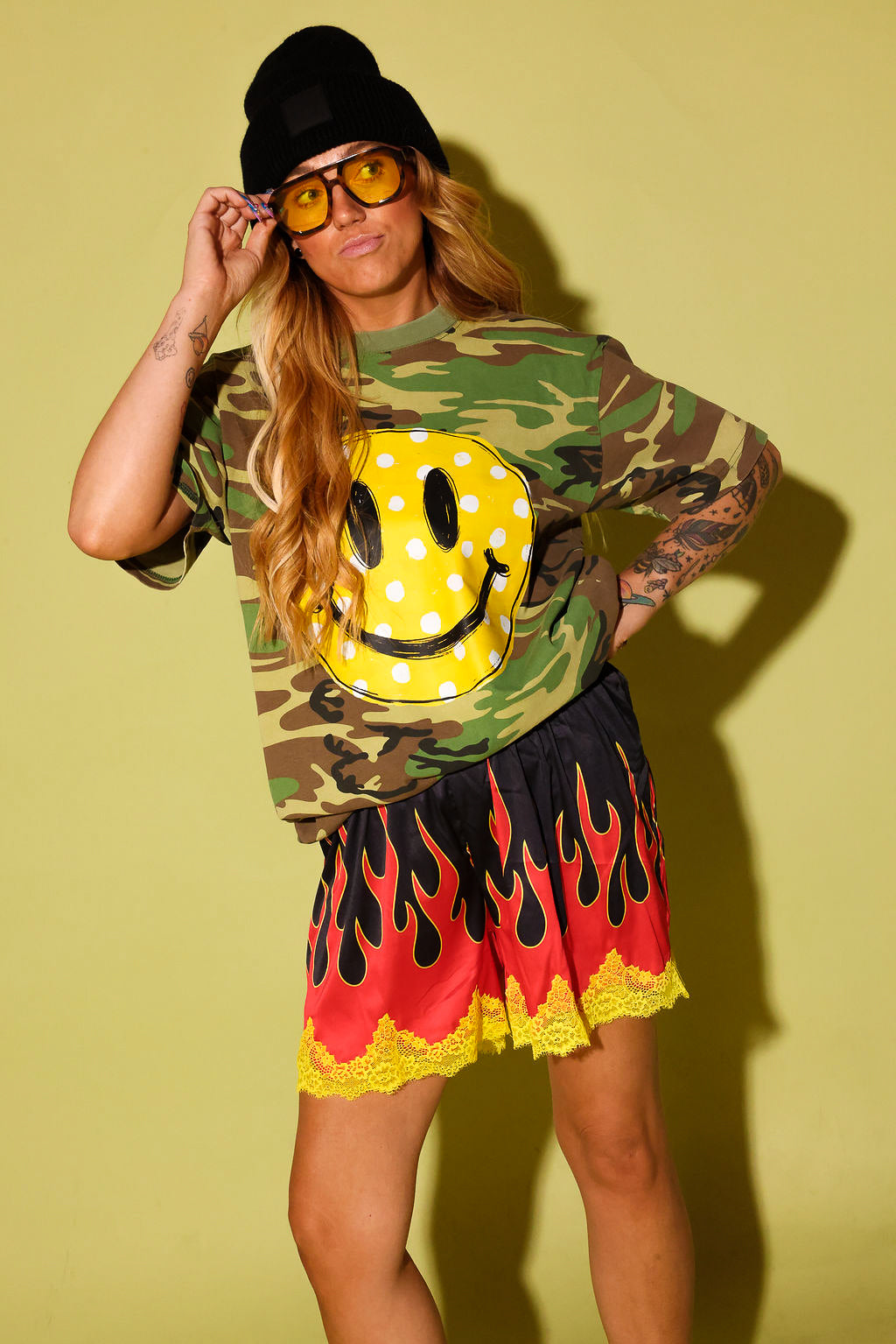 A woman modeling a camouflage long-sleeve t-shirt with a large yellow smiley face patch on the front, paired with a red and black flame-patterned skirt with yellow lace trim, a black beanie, and yellow-tinted sunglasses, set against a solid olive-green background.