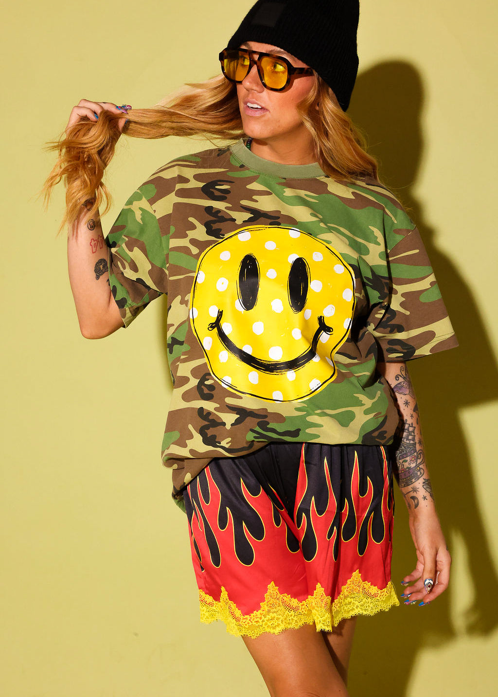 A woman modeling a green camouflage t-shirt with a large yellow smiley face graphic, paired with red and yellow flame-patterned shorts, a black beanie, and yellow-tinted sunglasses, set against a solid yellow background.
