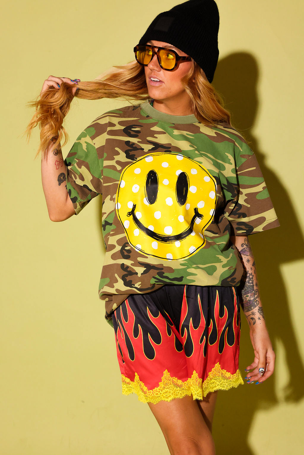 A woman modeling a green camouflage t-shirt with a large yellow smiley face graphic, paired with red and yellow flame-patterned shorts, a black beanie, and yellow-tinted sunglasses, set against a solid yellow background.