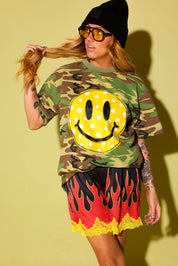 A woman modeling a green camouflage t-shirt with a large yellow smiley face graphic, paired with red and yellow flame-patterned shorts, a black beanie, and yellow-tinted sunglasses, set against a solid yellow background.