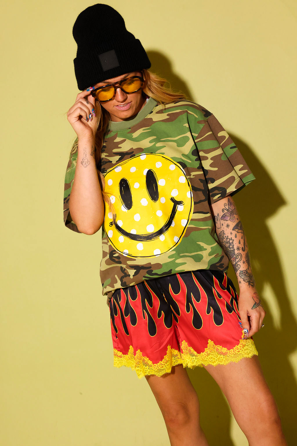 A model wearing a green camouflage t-shirt with a large yellow smiley face graphic, paired with red and black flame-patterned shorts with yellow lace trim, a black beanie, and yellow sunglasses, set against a solid yellow background.