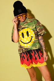 A model wearing a green camouflage t-shirt with a large yellow smiley face graphic, paired with red and black flame-patterned shorts with yellow lace trim, a black beanie, and yellow sunglasses, set against a solid yellow background.