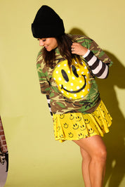 A woman modeling a camouflage long-sleeve top with a large yellow smiley face graphic on the front, paired with a yellow smiley-print pleated skirt and a black beanie, against a pale yellow-green studio background.