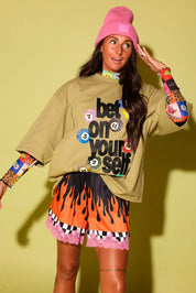 A woman modeling a beige graphic t-shirt with 'best on your self' text and colorful circular accents, paired with orange and black flame-patterned shorts with pink lace trim, a pink beanie, and colorful patterned arm sleeves, set against a solid yellow-green studio background.