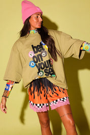 A woman modeling an oversized beige graphic sweatshirt with colorful smiley face and number prints, paired with orange and pink patterned shorts, a pink beanie, and patterned long sleeves, posed against a solid yellow-green studio background.
