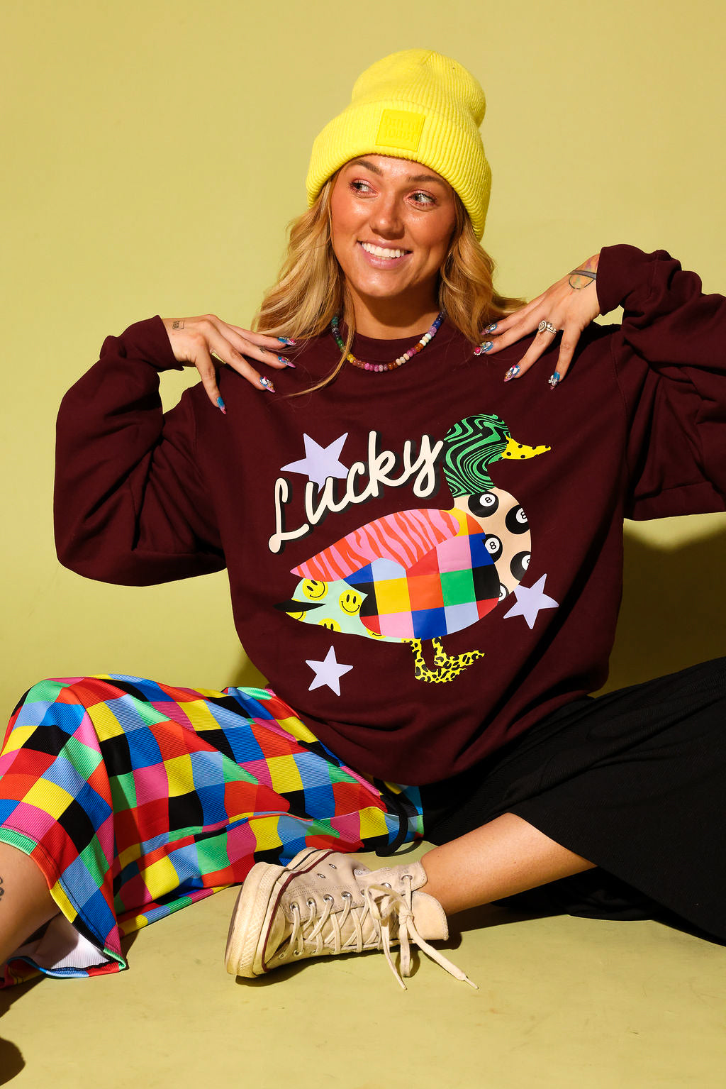 A woman wearing a burgundy sweatshirt with a colorful fish graphic and the word 'Lucky', paired with a yellow beanie, a multicolor checkered skirt, and beige sneakers, posing against a solid yellow-green background.