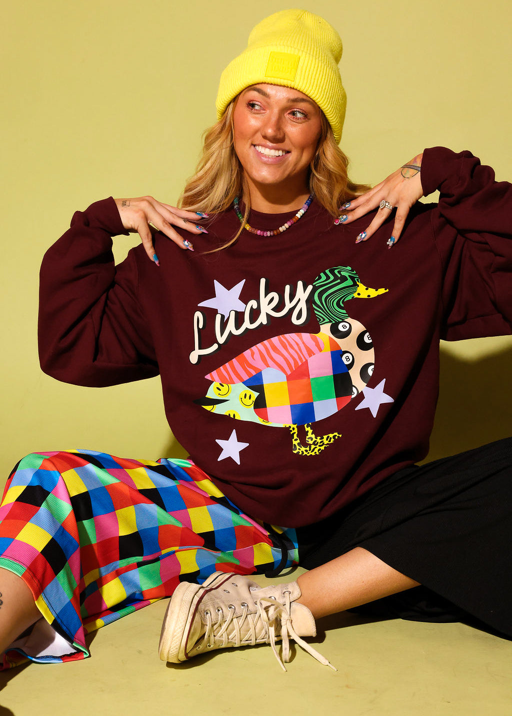 A woman wearing a burgundy sweatshirt with a colorful fish graphic and the word 'Lucky', paired with a yellow beanie, a multicolor checkered skirt, and beige sneakers, posing against a solid yellow-green background.