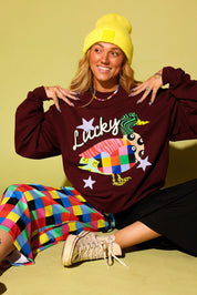 A woman wearing a burgundy sweatshirt with a colorful fish graphic and the word 'Lucky', paired with a yellow beanie, a multicolor checkered skirt, and beige sneakers, posing against a solid yellow-green background.