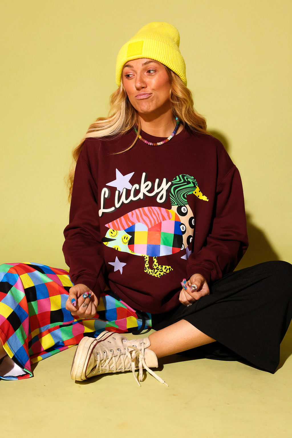 A woman sits cross-legged against a solid pale yellow-green background, wearing a maroon sweatshirt with a colorful duck graphic and the word 'Lucky', a bright yellow beanie, a multicolor checkered skirt, and beige sneakers.