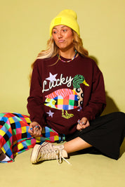 A woman sits cross-legged against a solid pale yellow-green background, wearing a maroon sweatshirt with a colorful duck graphic and the word 'Lucky', a bright yellow beanie, a multicolor checkered skirt, and beige sneakers.